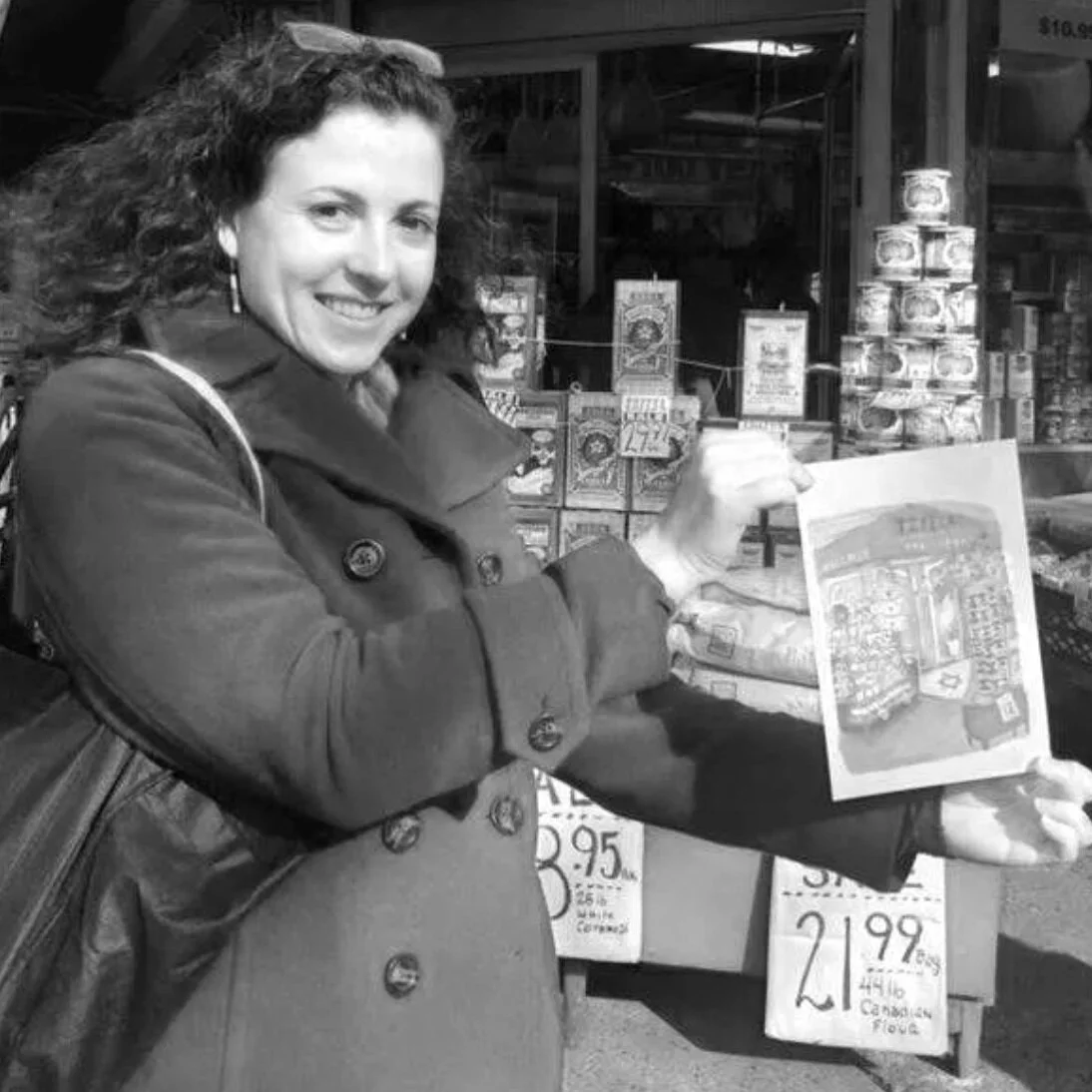 A woman with curly hair smiling and holding a paper or brochure in front of store display shelves with canned goods and handwritten sale signs.