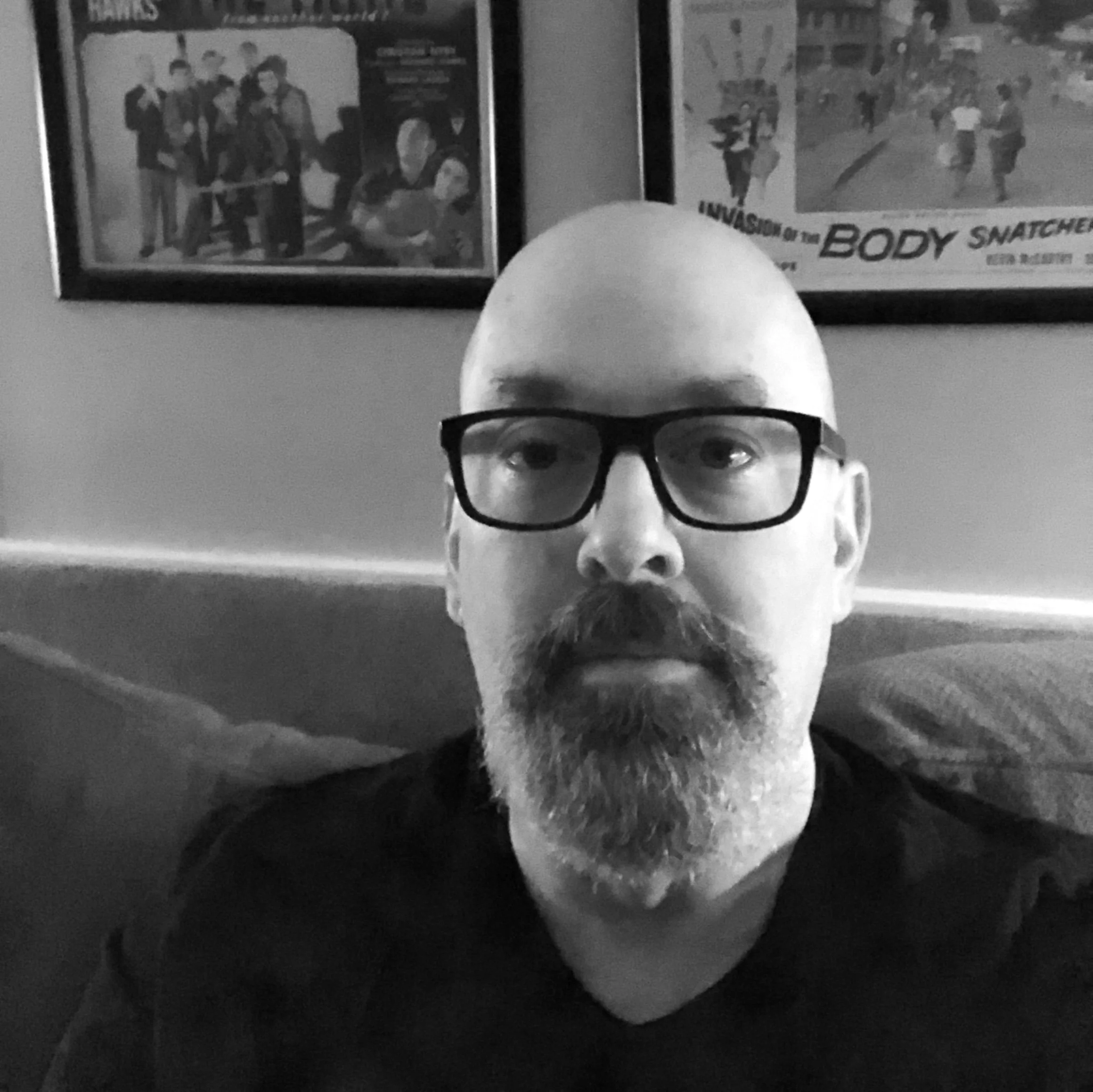 A man with glasses and a beard taking a selfie in a room with framed posters on the wall behind him.