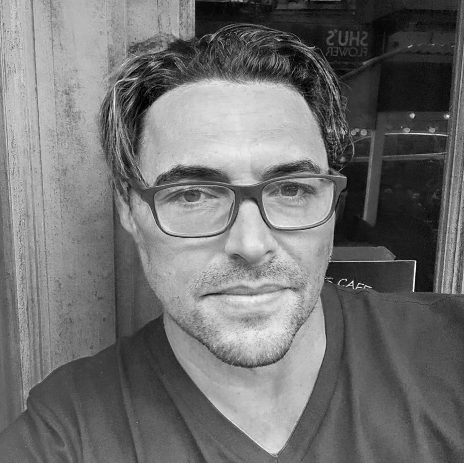 A black-and-white photo of a man with glasses and short, dark hair, sitting indoors near a wooden wall and window.