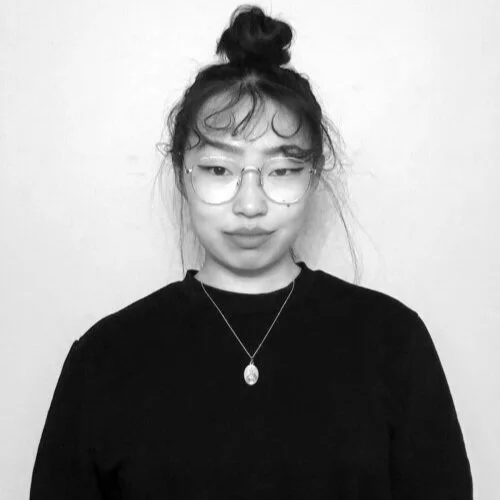 Black-and-white photo of a young woman with glasses, wearing a top knot hairstyle, a necklace, and a black shirt, standing against a plain background.