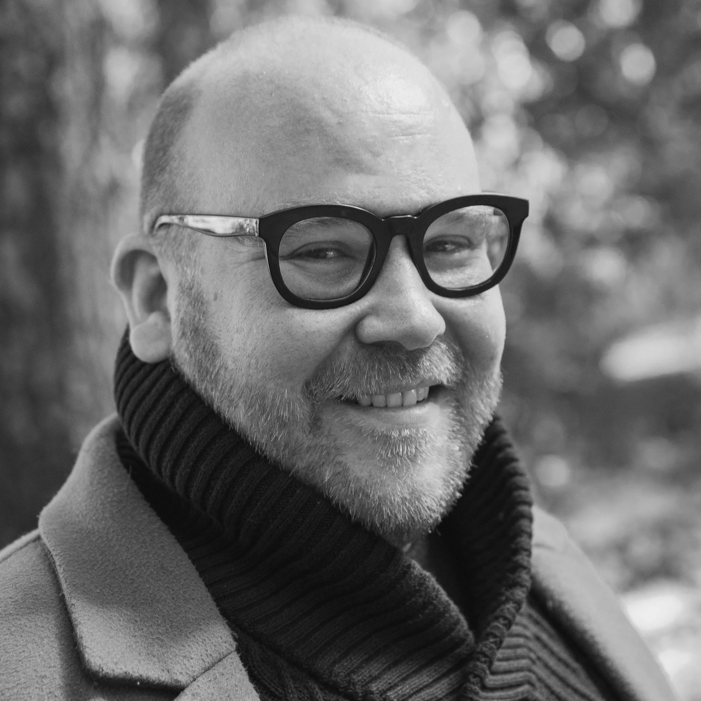A smiling man wearing glasses, a turtleneck sweater, and a coat outdoors, with a blurred background of trees.