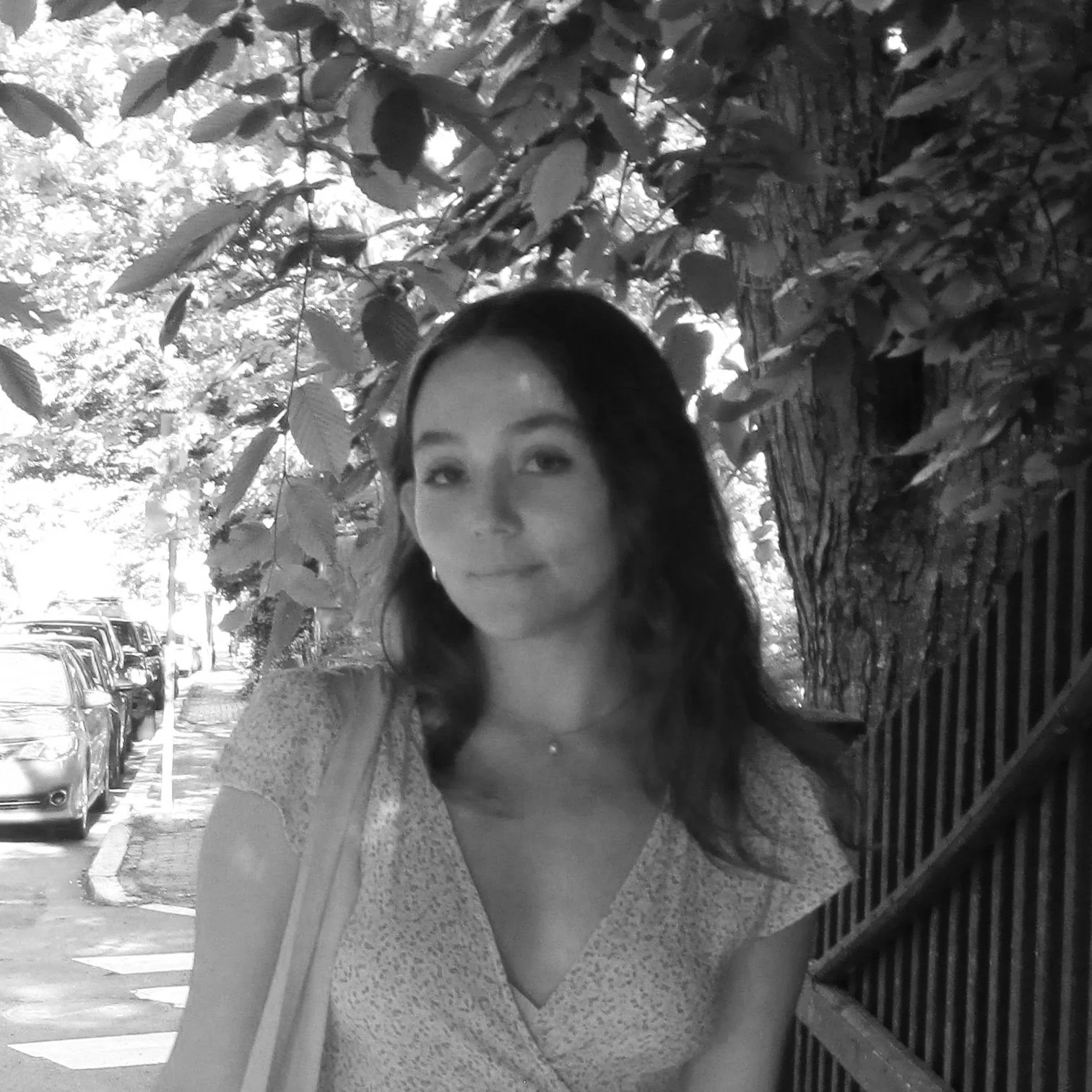A young woman with long dark hair standing outside on a sidewalk next to a tree, with parked cars and a fence in the background, captured in black and white.
