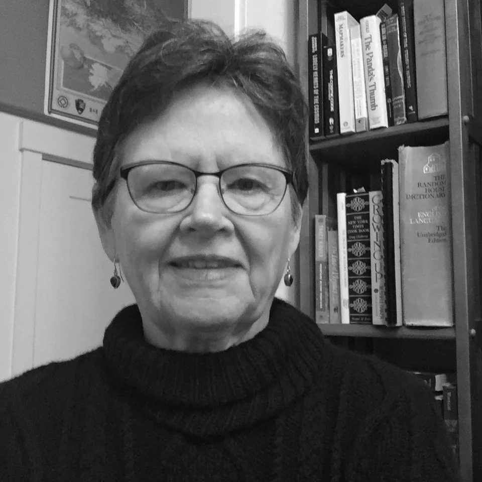 A black and white photo of an older woman with short hair, glasses, and earrings, standing in front of a bookshelf filled with books and a framed picture on the wall.