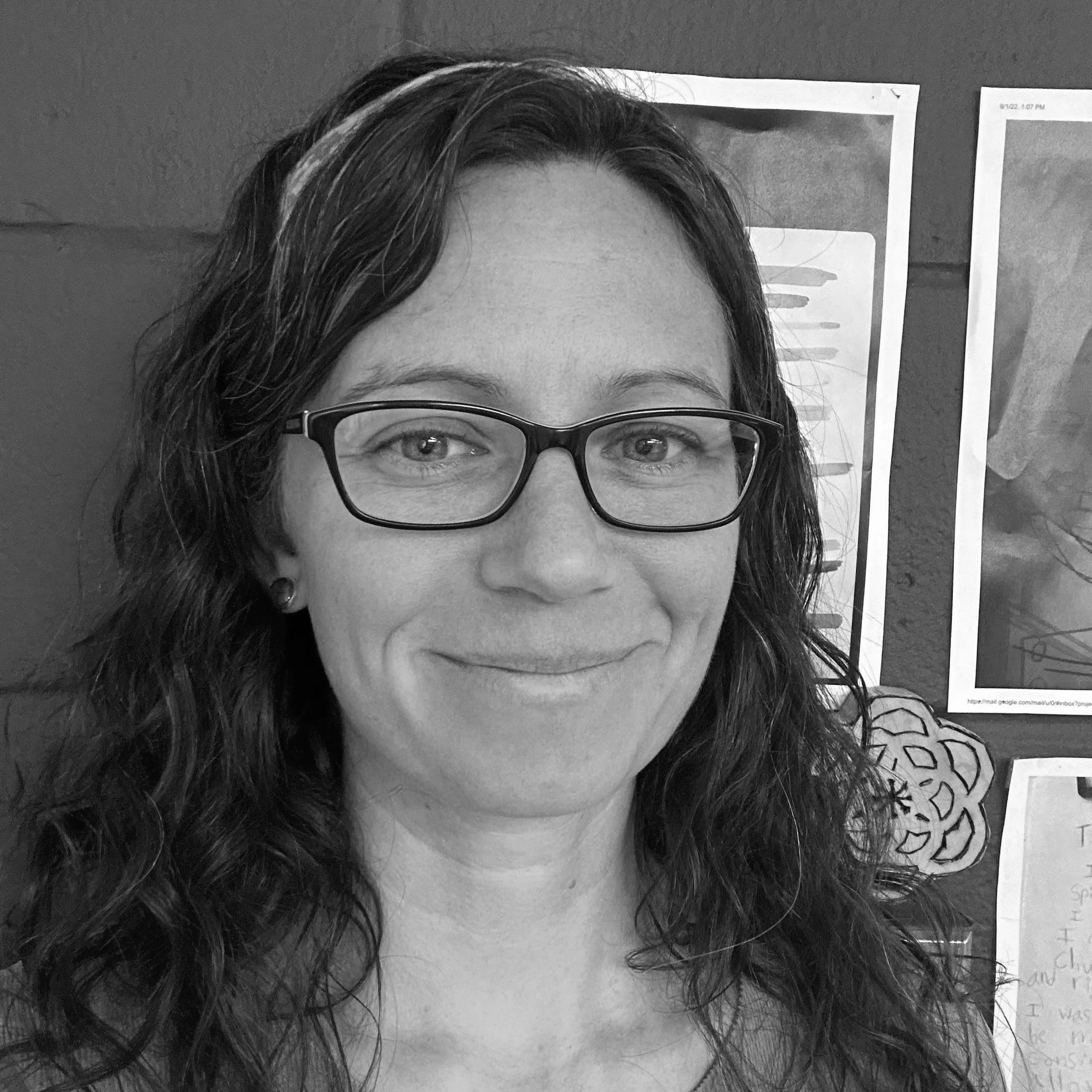 A woman with glasses and long wavy hair smiling in front of a bulletin board with photos and papers.