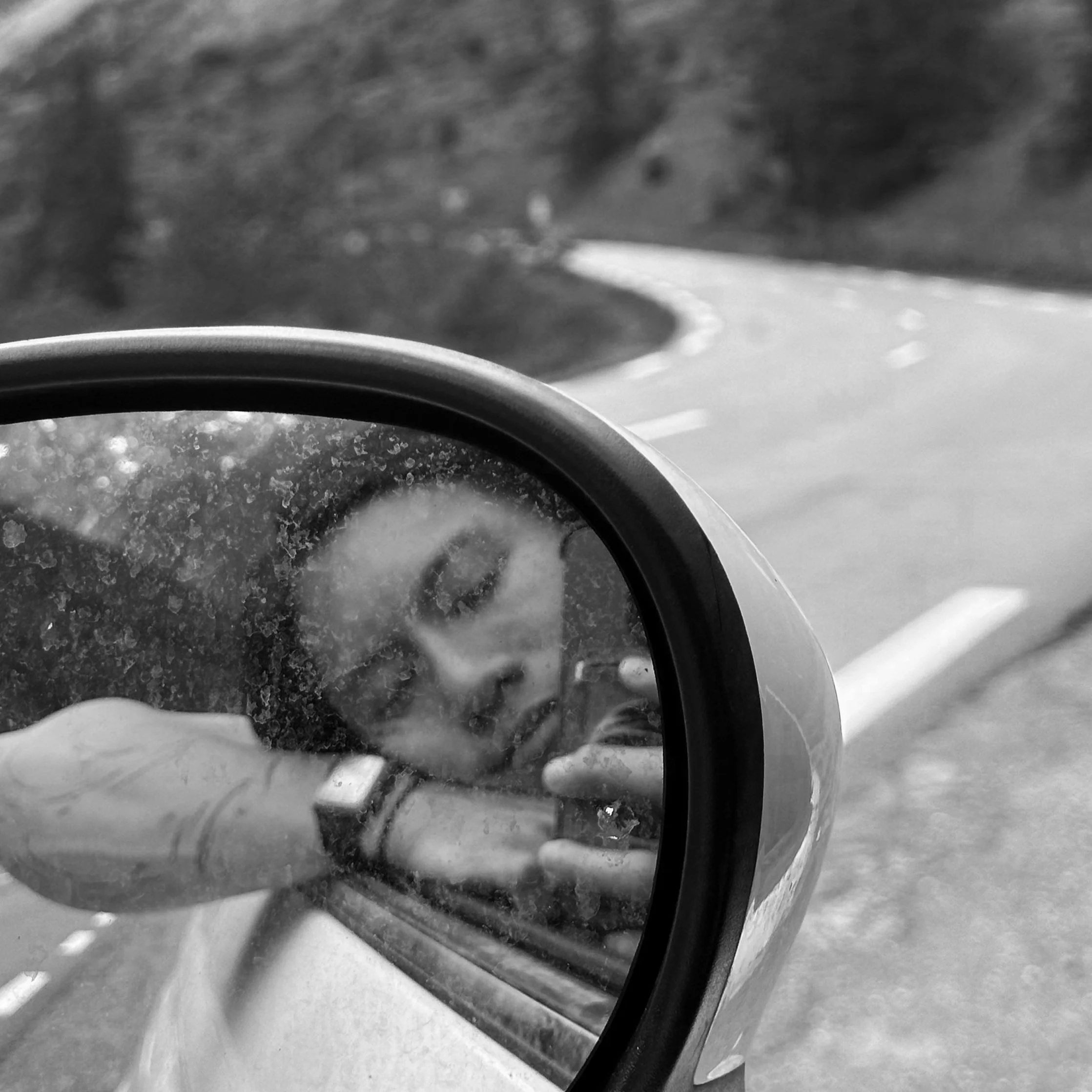 A person taking a selfie in a vehicle's side mirror, with a winding road and landscape in the background, in black and white.