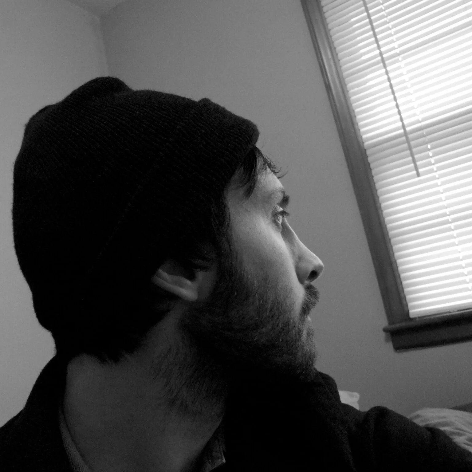 Black and white side profile of a man wearing a beanie, looking out a window with blinds.