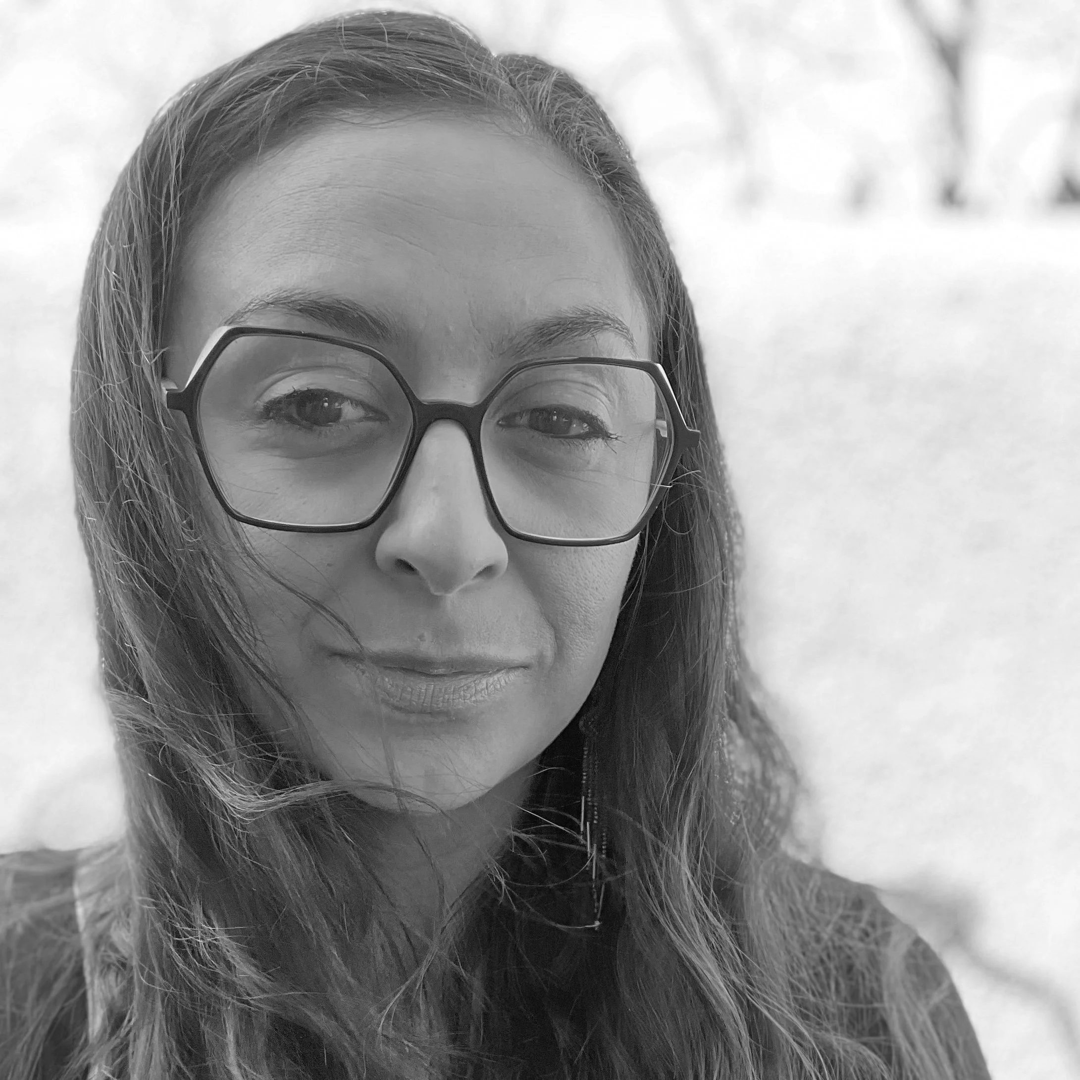 Black and white photo of a woman with glasses, long wavy hair, and a slight smile, standing outdoors.