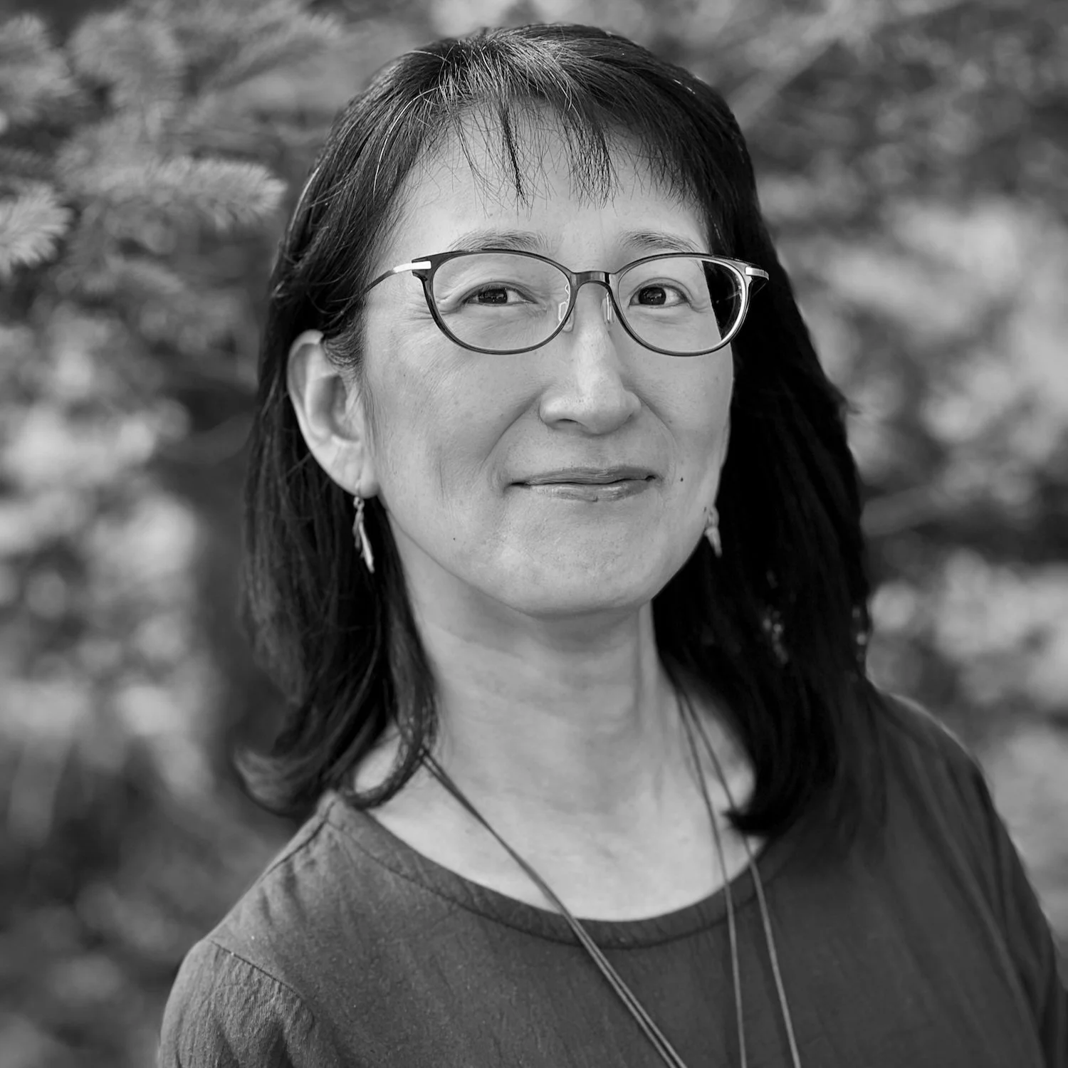 Portrait of an Asian woman with glasses, dark hair, earrings, and a necklace, outdoors with blurred natural background.