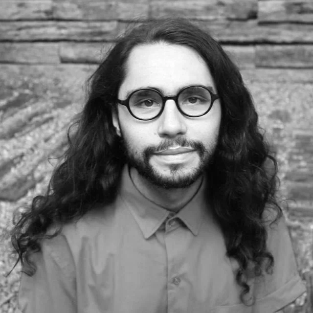 Black and white portrait of a man with long curly hair, glasses, and a beard, wearing a collared shirt, standing outdoors against a wooden background.