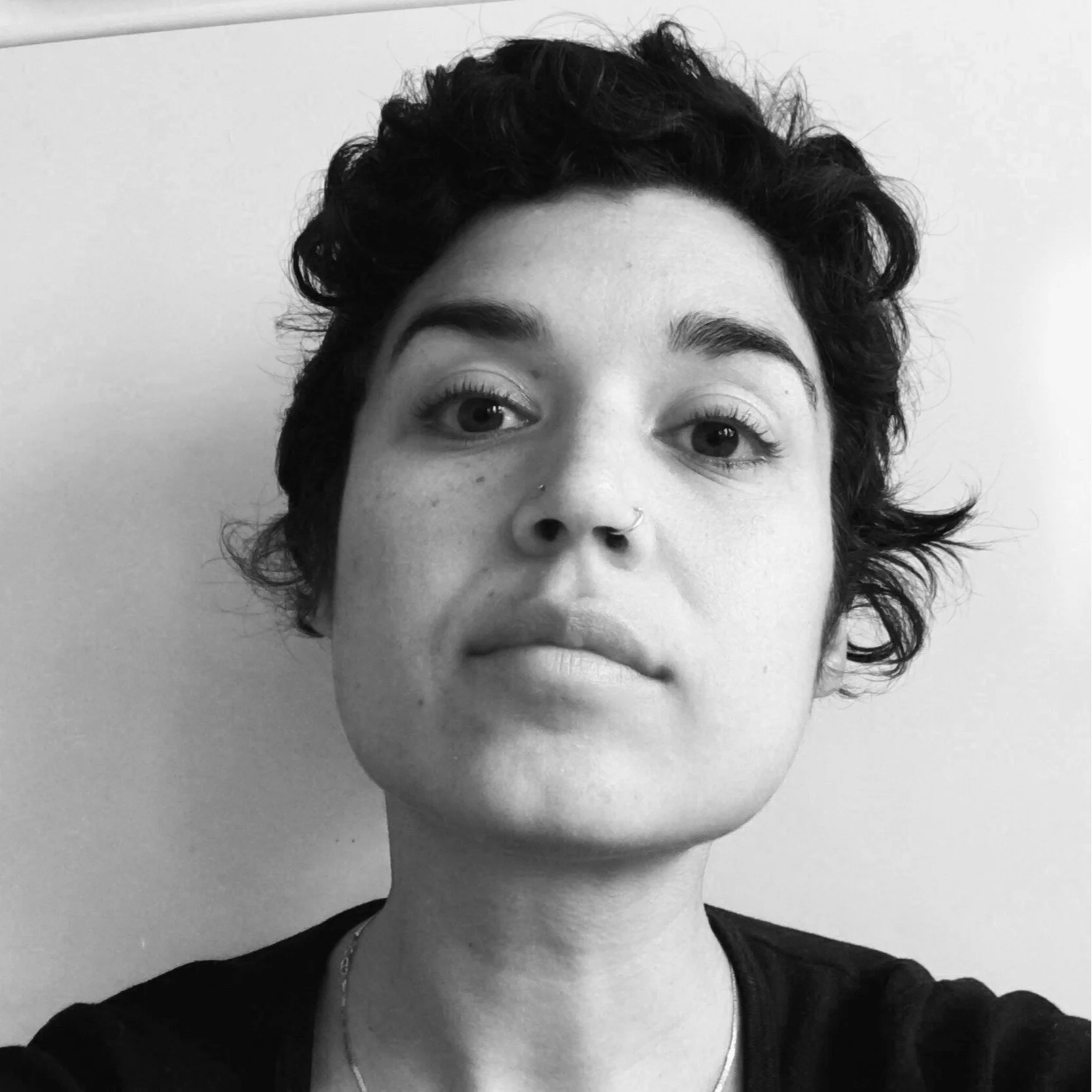 A black and white close-up portrait of a woman with short, curly hair, wearing a nose ring and a necklace, looking directly at the camera.