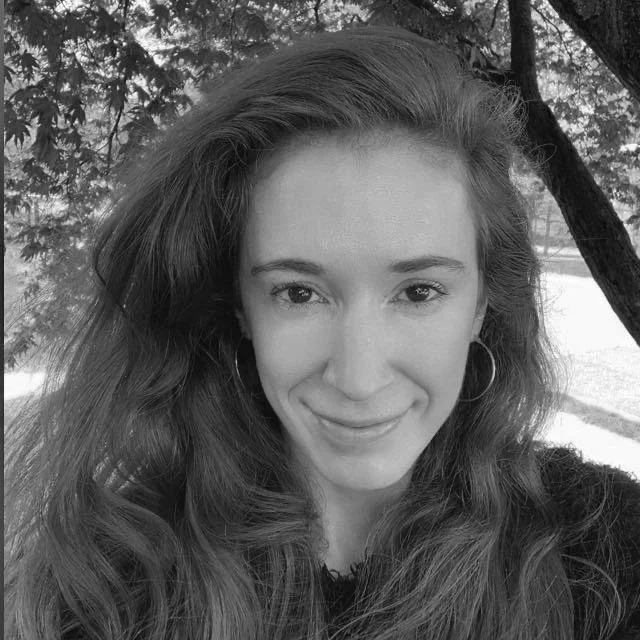 A young woman with long, wavy hair smiling outdoors in front of trees, in black and white.