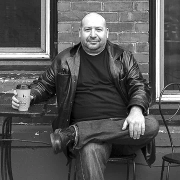 Man sitting outside, holding a coffee cup, wearing a leather jacket, in front of a brick wall with a window.