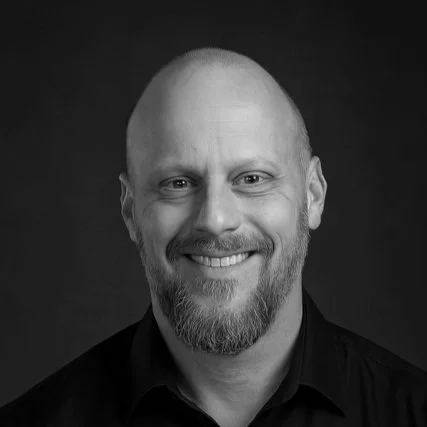 Black and white portrait of a smiling man with a beard and shaved head, wearing a dark shirt.