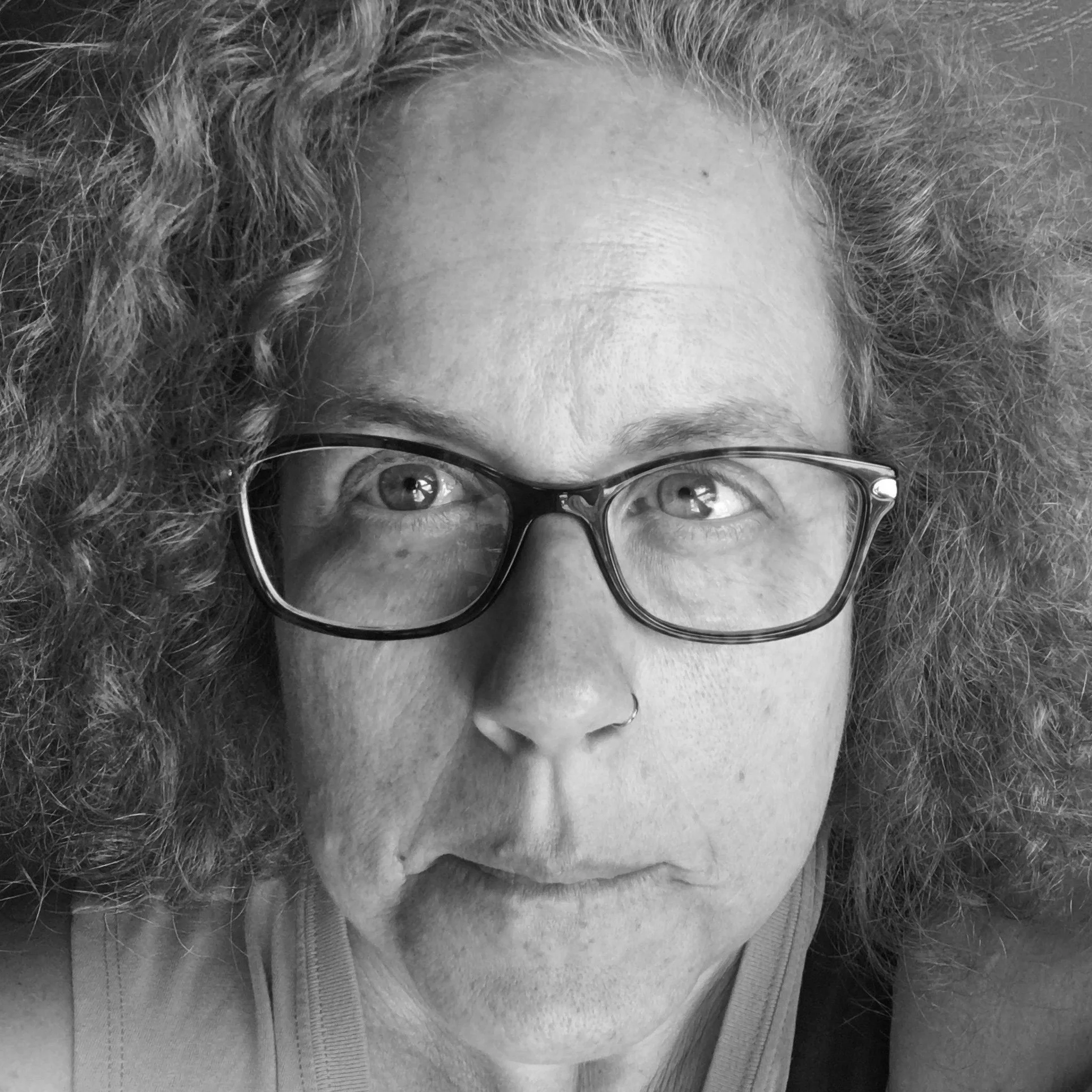 Close-up black-and-white photo of a woman with curly hair wearing glasses and a nose ring, looking slightly upward.