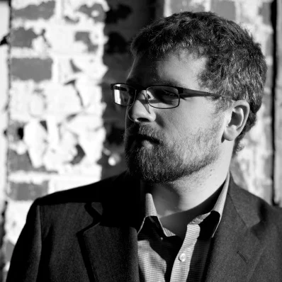 A man with glasses and a beard looks to his left against a brick wall background.