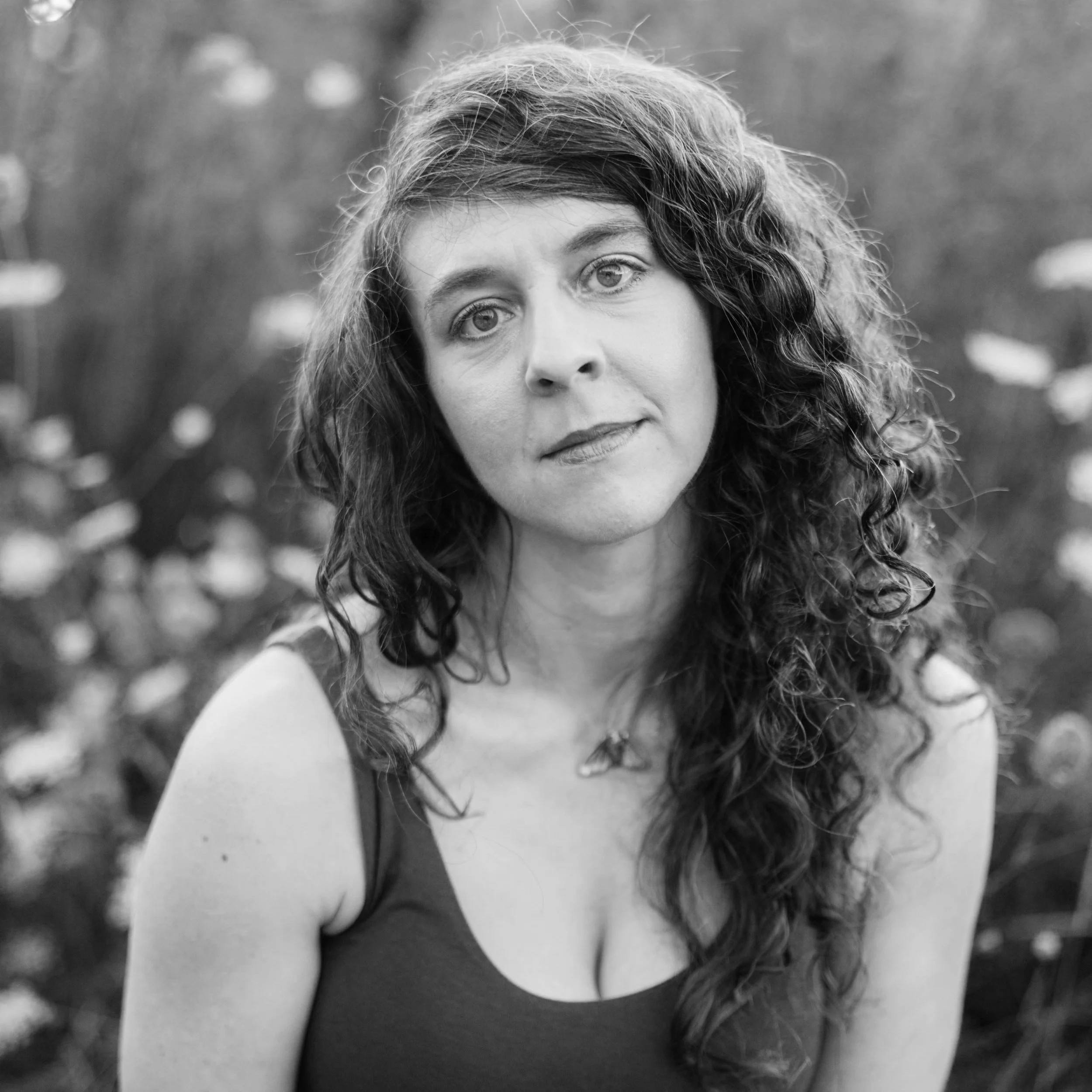 Black and white portrait of a woman with long curly hair, wearing a sleeveless top and a necklace, outdoors with blurred foliage in the background.
