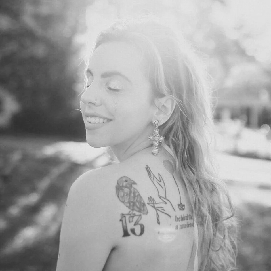 Black and white photo of a smiling woman with closed eyes, earrings, and a tattoo on her shoulder, outdoors.