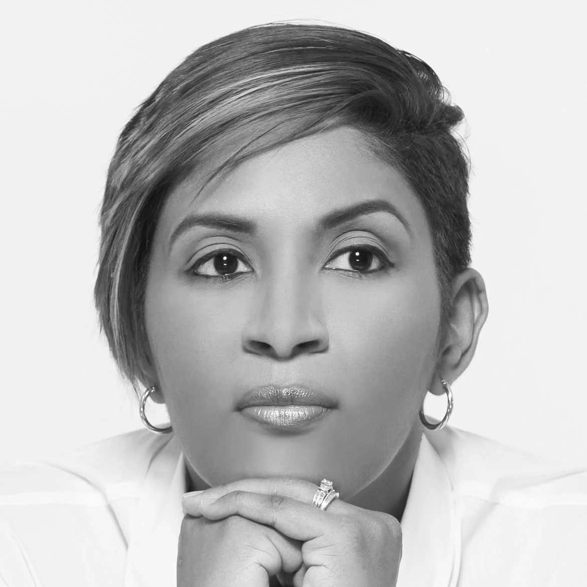 Black and white portrait of a woman with short, styled hair, wearing hoop earrings, resting her chin on her hand with rings, looking directly at the camera.