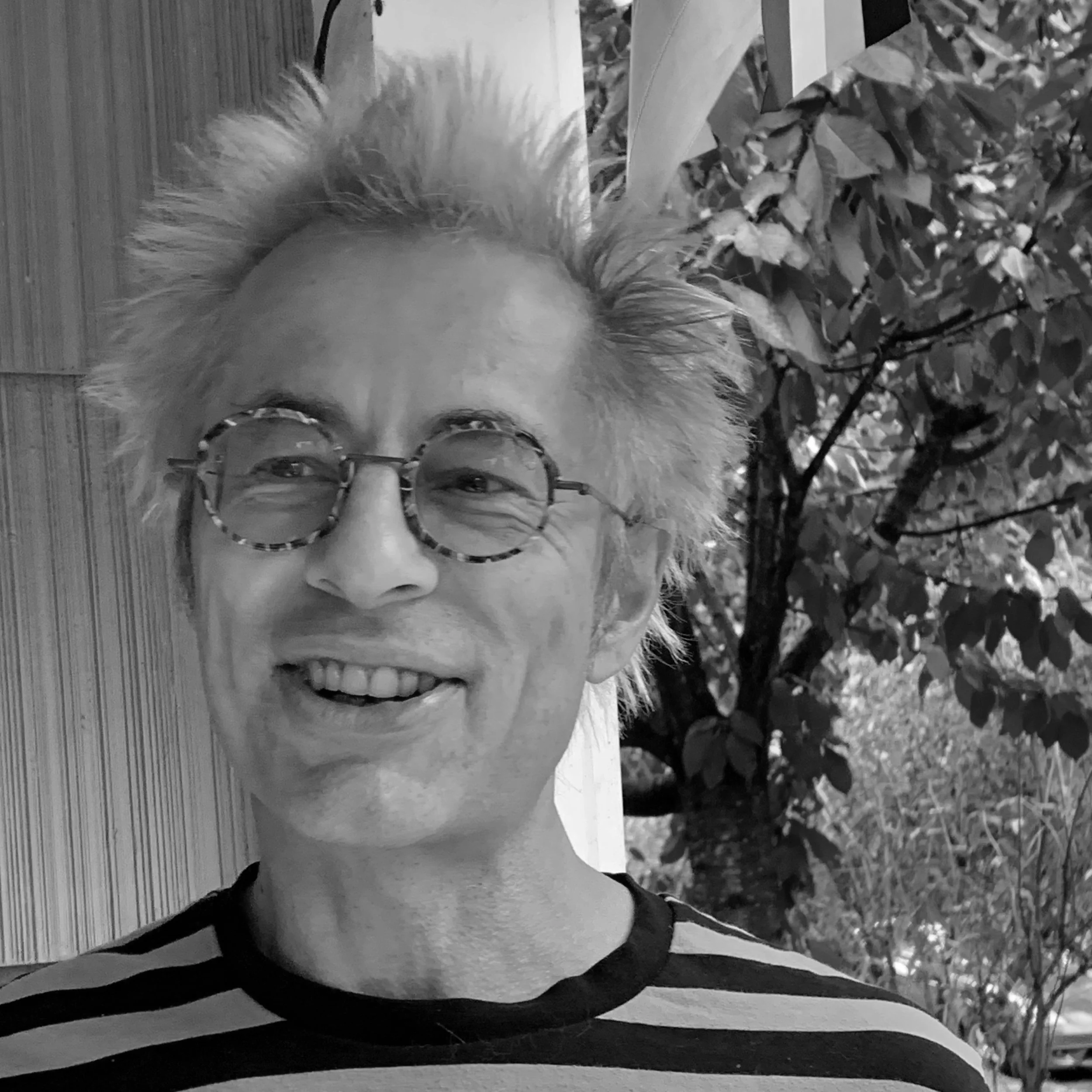 A smiling man with glasses, spiky hair, and a striped shirt, standing outdoors with trees and foliage in the background.