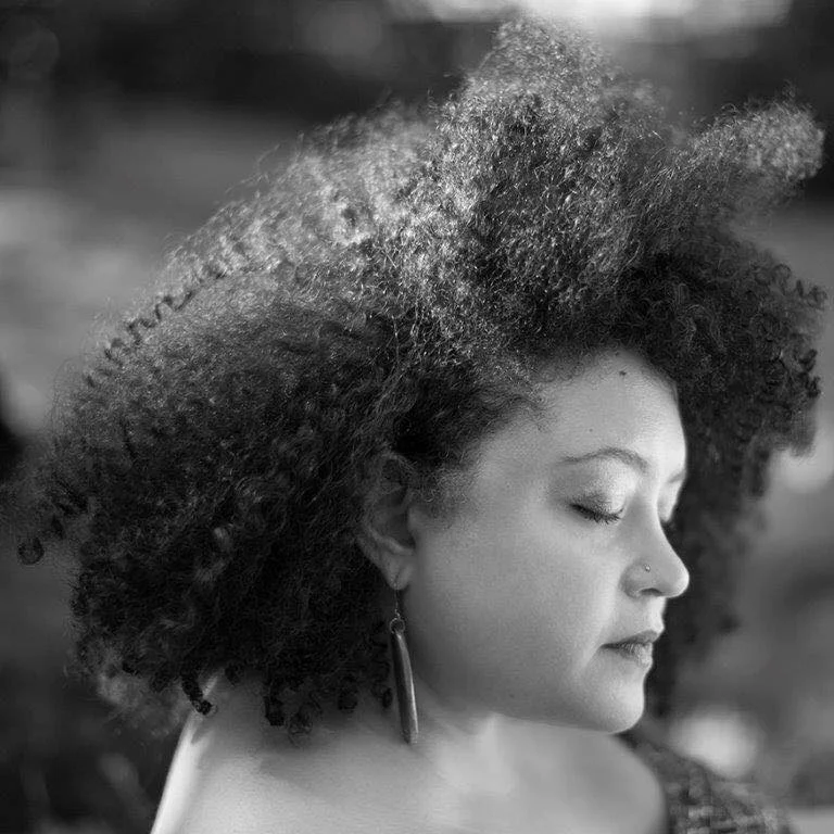 A woman with natural curly hair and a nose piercing, in profile view with her eyes closed.