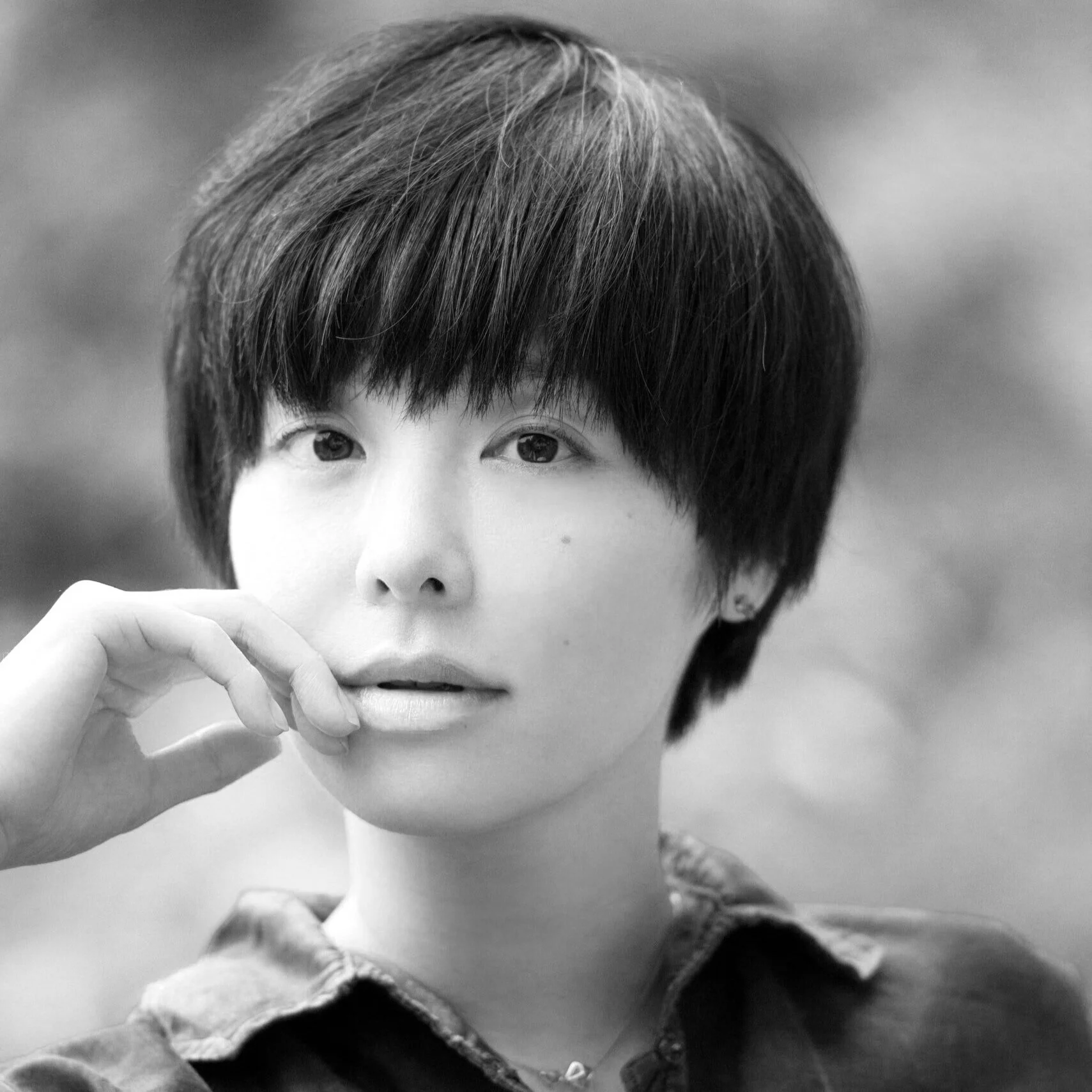 Black and white portrait of a woman with short, layered hair, gently touching her lips with her fingers, in an outdoor setting.
