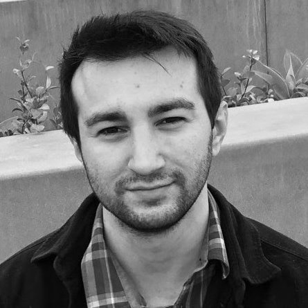 A young man with short dark hair and light facial hair, wearing a checkered shirt and dark jacket, smiling slightly outdoors near plants.