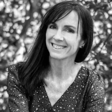 A woman with dark hair smiling outdoors with a blurred background of trees