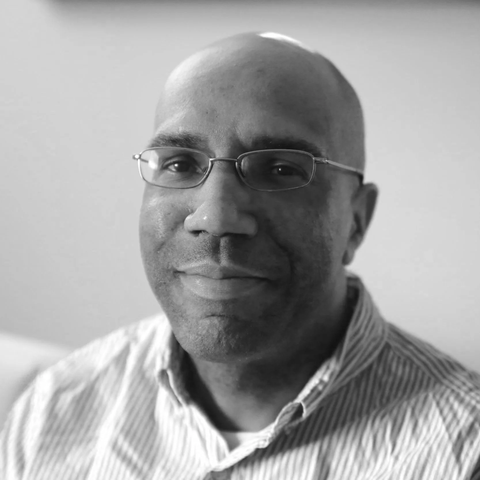 Black and white portrait of a bald man wearing glasses and a striped collared shirt, smiling subtly.