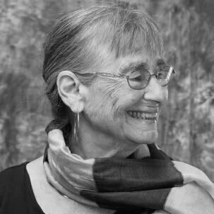 Profile of an elderly woman with short hair and glasses, smiling, outdoors in a natural setting.