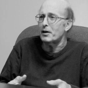 A black-and-white photo of an older man with glasses, speaking or explaining something while sitting in an armchair.