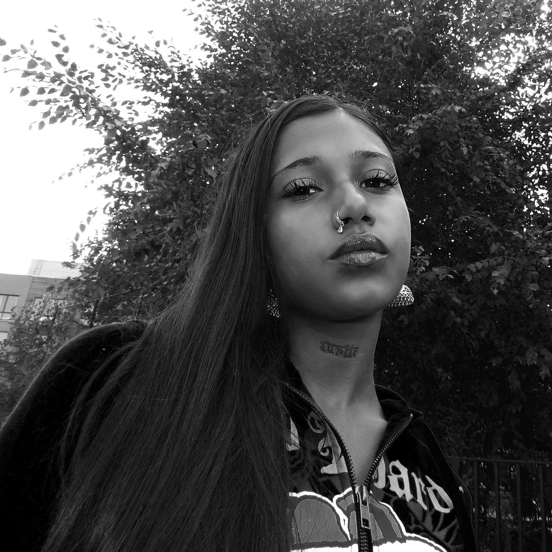 A young woman with long straight hair, wearing large earrings and a nose ring, looking confidently at the camera outdoors with trees in the background.