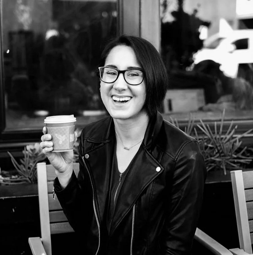 A smiling woman with short dark hair and glasses holding a to-go coffee cup, sitting outdoors near a window.