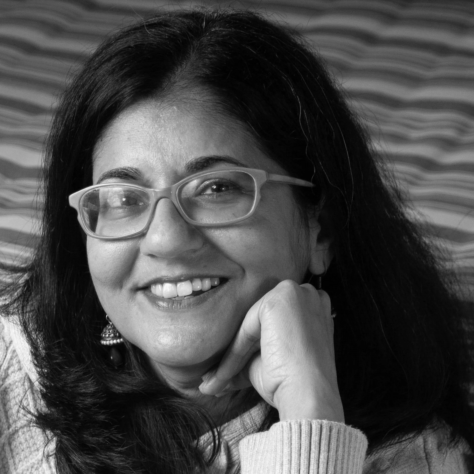A woman with glasses smiling and resting her chin on her hand, with wavy hair and a patterned background.