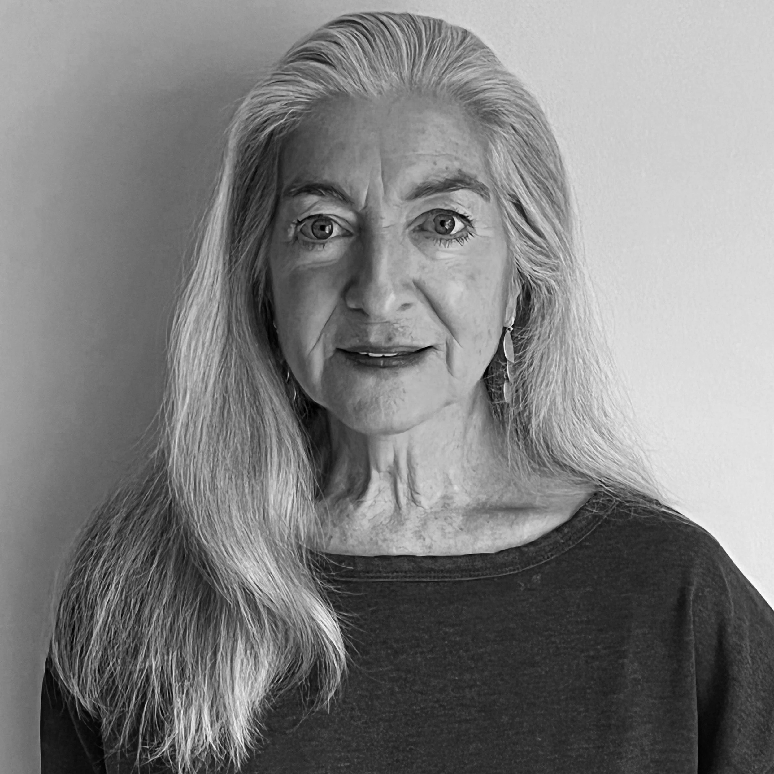 Black and white portrait of an older woman with long, straight hair, wearing a dark top and earrings, looking directly at the camera.