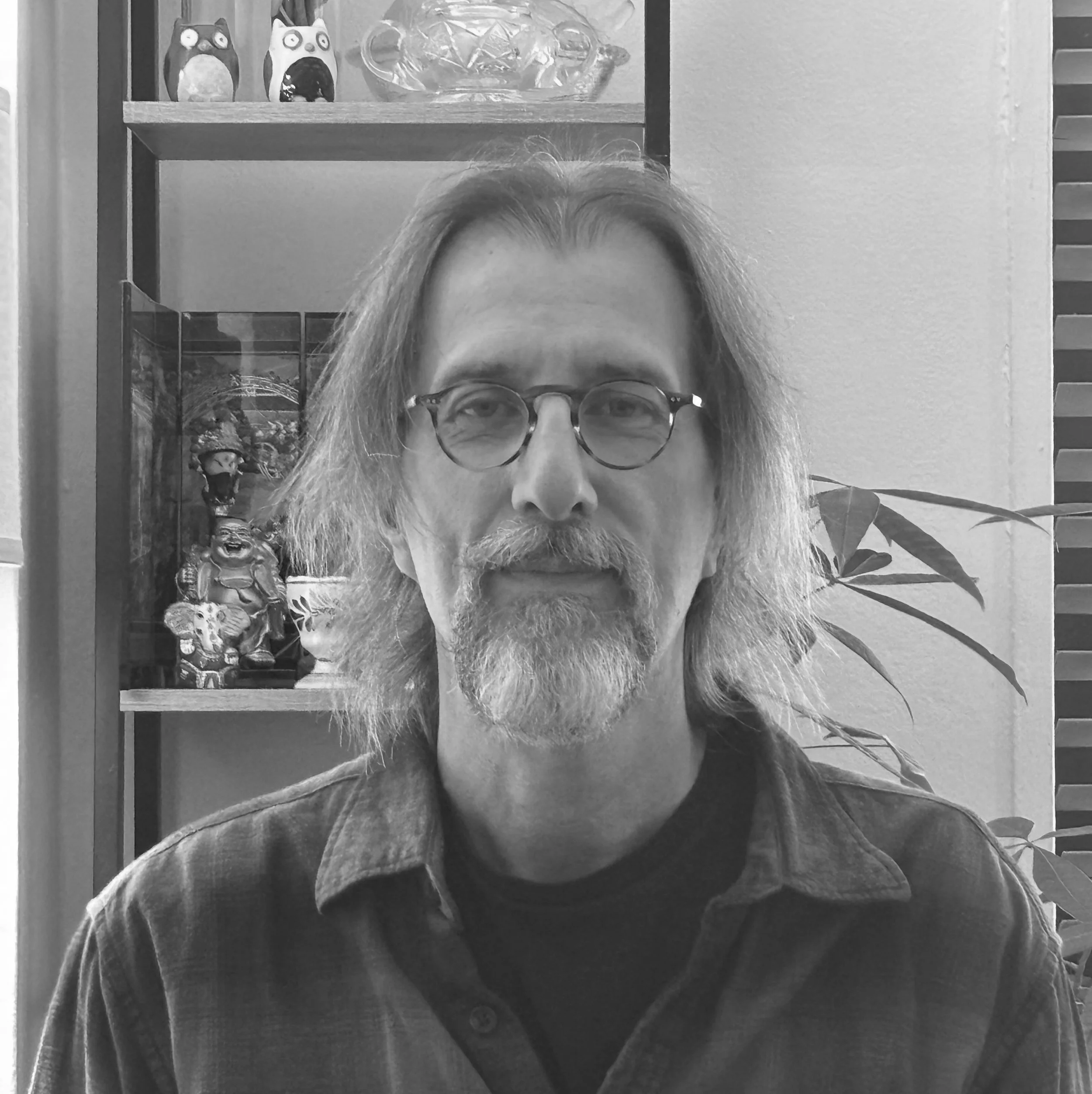 Black and white portrait of a man with shoulder-length hair, beard, glasses, wearing a plaid shirt, sitting in front of a shelf with decorative items and a plant.