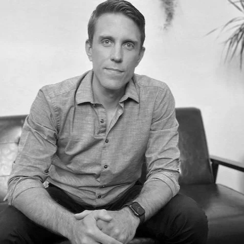 A man with short hair and light skin sitting on a chair, wearing a button-up shirt and a watch, with a serious expression in a black and white photo.