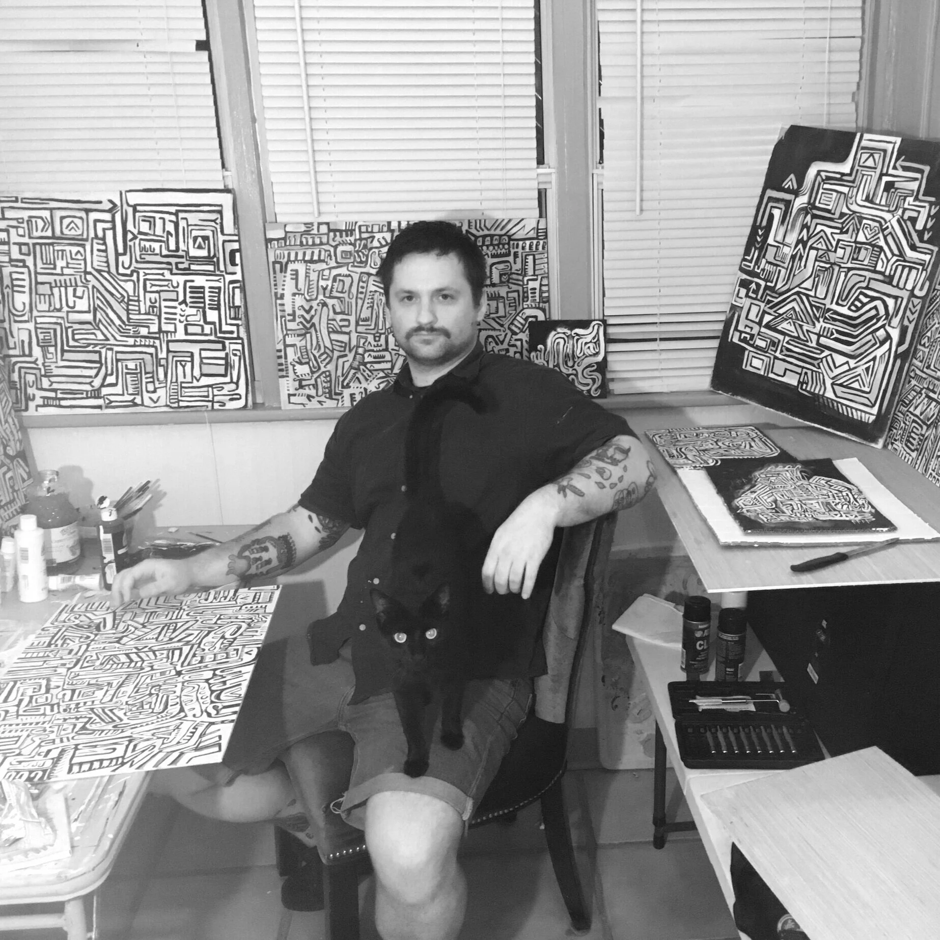 A man with tattoos on his arms sitting at a table with paintings of abstract black and white doodles behind him. He's holding a black cat on his lap, and the room has a window with blinds.