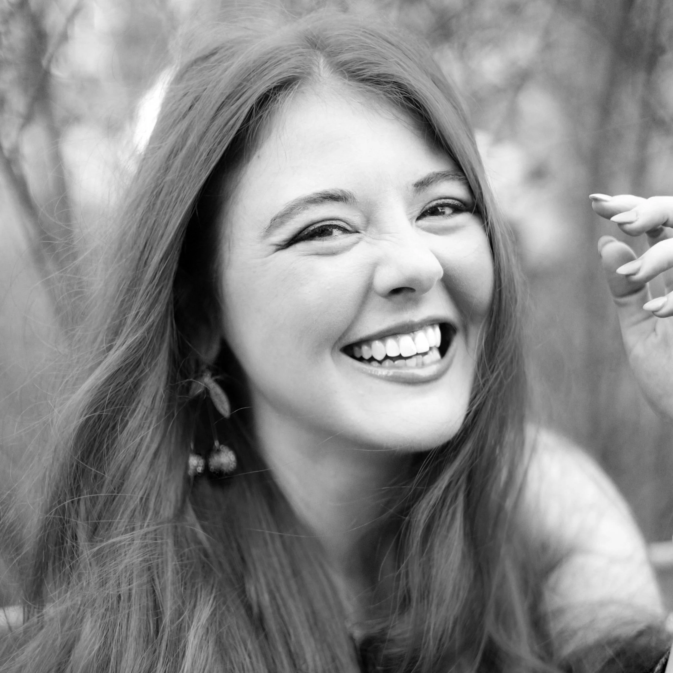 Black and white photo of a woman with long hair smiling and showing teeth, with one eye slightly squinted, outdoors with blurred background.