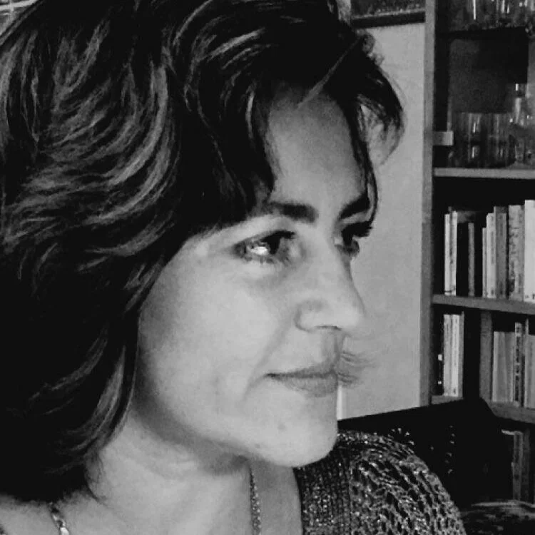 Black and white photo of a woman with short, wavy hair in a room with a bookshelf in the background.