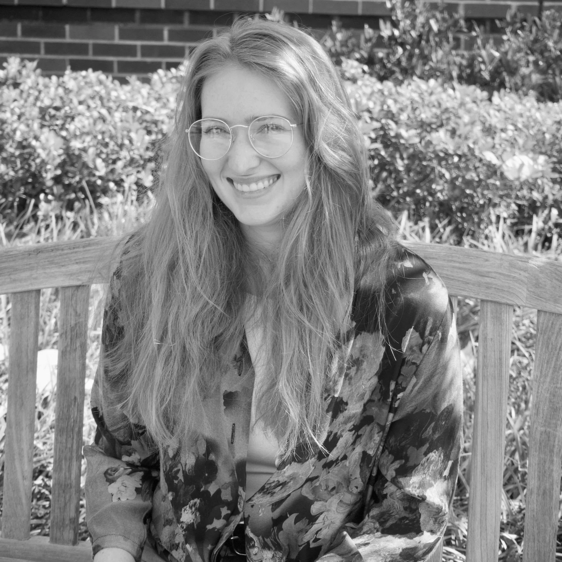 A woman with long wavy hair and glasses smiling outdoors, sitting on a wooden bench in front of a garden with flowers and a brick wall.