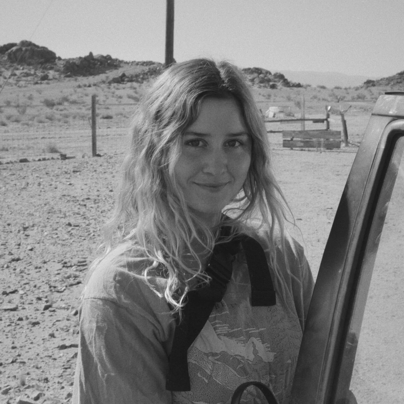 A woman with wavy hair smiling outdoors in a desert landscape, standing near a vehicle.