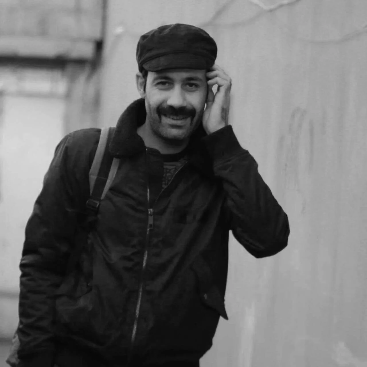 A man with a mustache and beard, wearing a dark cap and jacket, is smiling and holding his head with one hand.