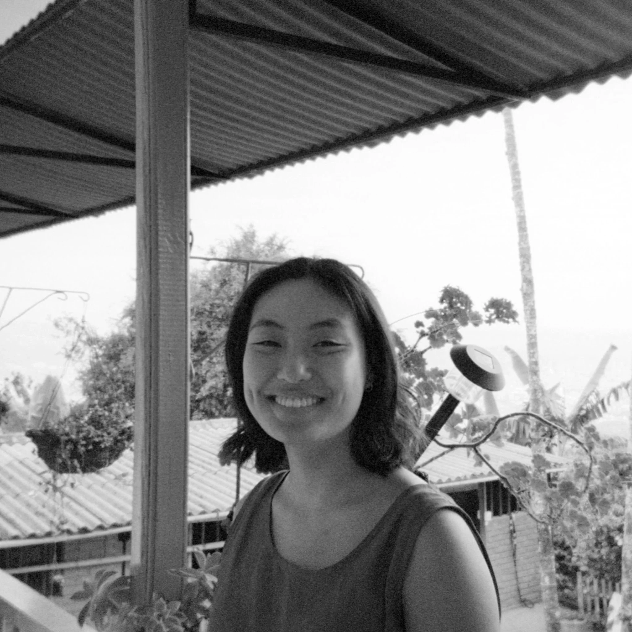 A smiling woman with shoulder-length hair standing on a porch, with a scooter and trees in the background, captured in black and white.