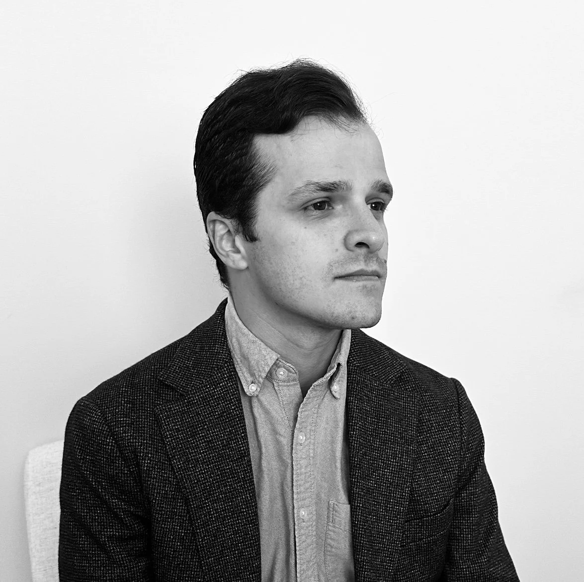 Black and white photo of a young man with dark hair, wearing a blazer and button-up shirt, sitting against a plain light-colored background.