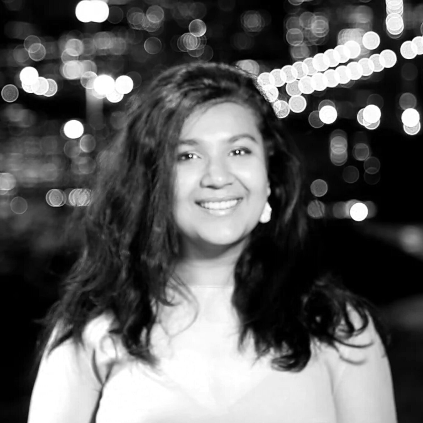 A woman with long dark hair smiling at night with city lights and bokeh effects in the background.