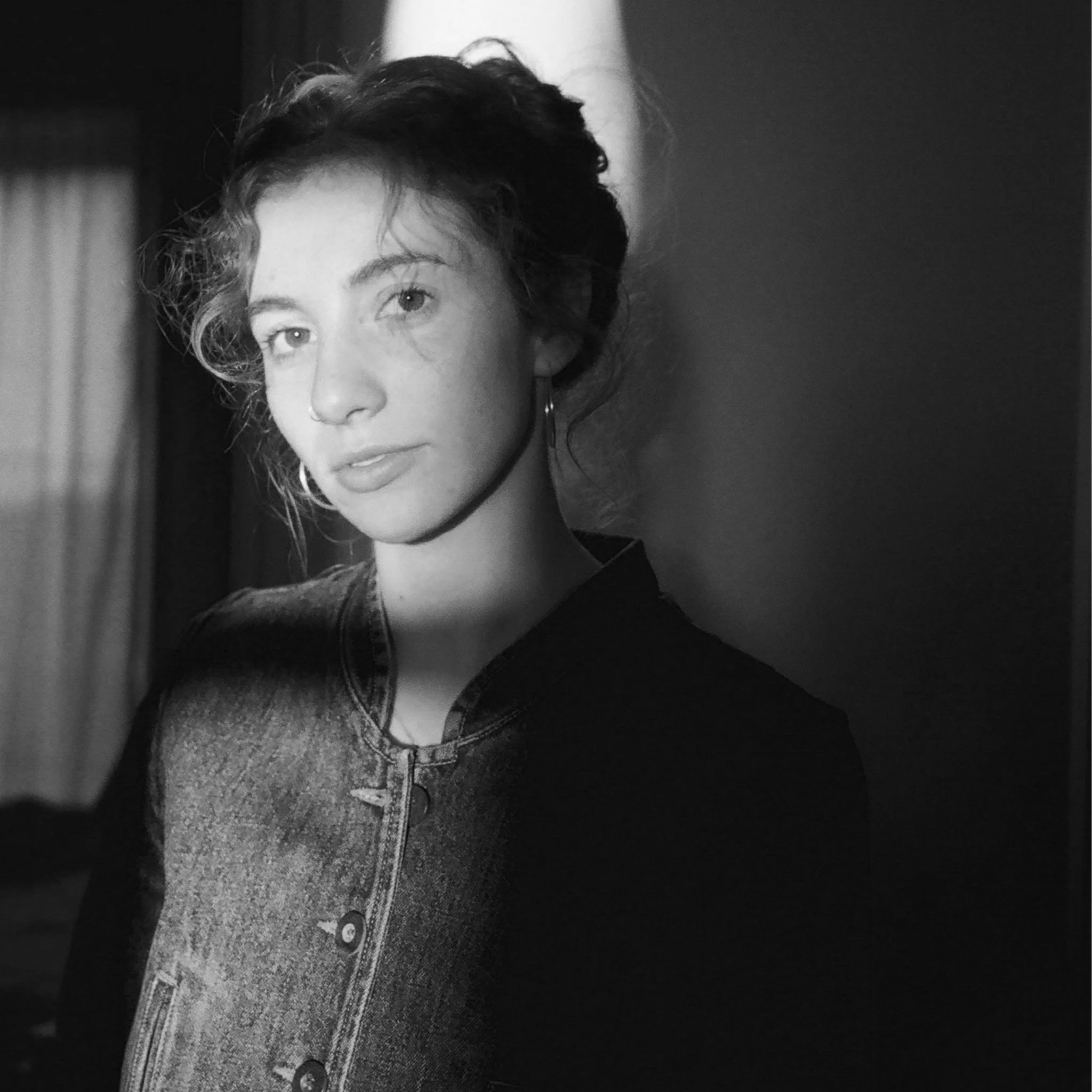 Black and white portrait of a woman with short curly hair, looking at the camera with a soft expression, wearing a denim jacket, in a room with curtains in the background.