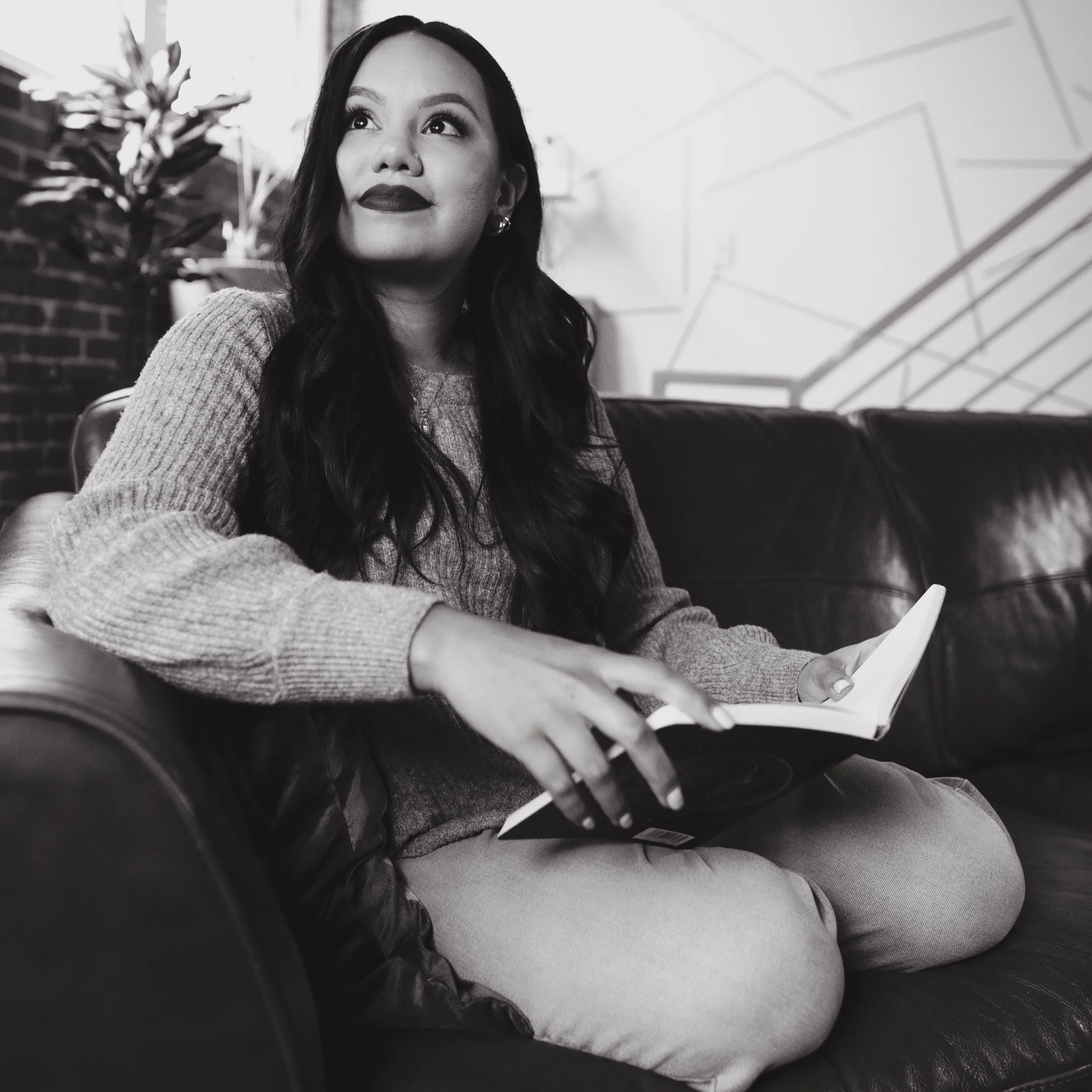 A woman sitting on a leather couch, holding an open book, with long dark hair, wearing a sweater, in a modern indoor setting.