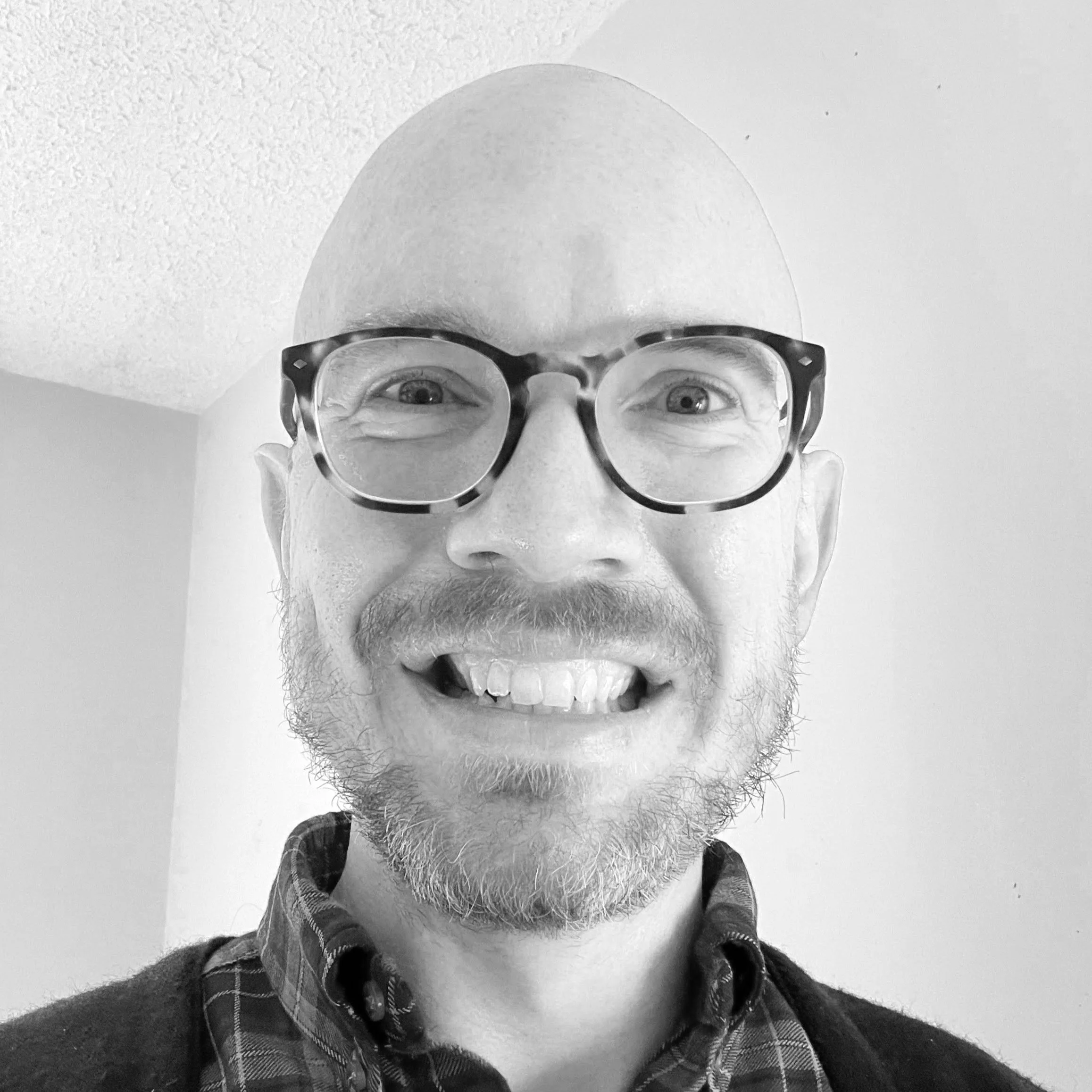 A smiling man with glasses, a beard, and a missing front tooth, wearing a checkered shirt and a dark sweater, indoors with a plain wall and ceiling in the background.