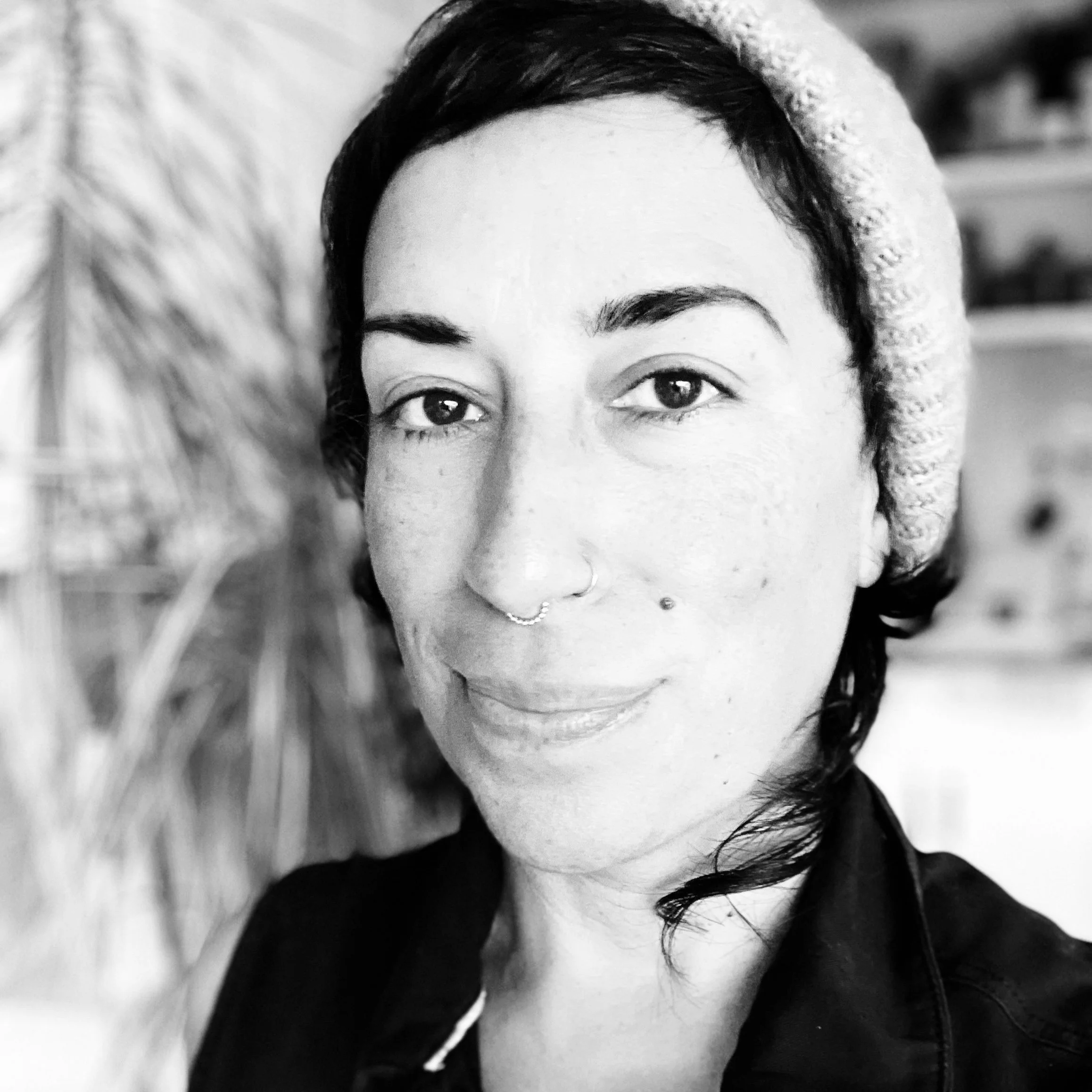 A woman with short dark hair, wearing a knitted hat with a pom-pom, smiling slightly, with a plant in the background.