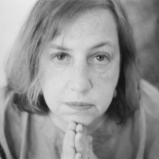 Close-up black and white photograph of a woman with short hair, looking directly at the camera with a thoughtful expression, her hands pressed together near her chin.